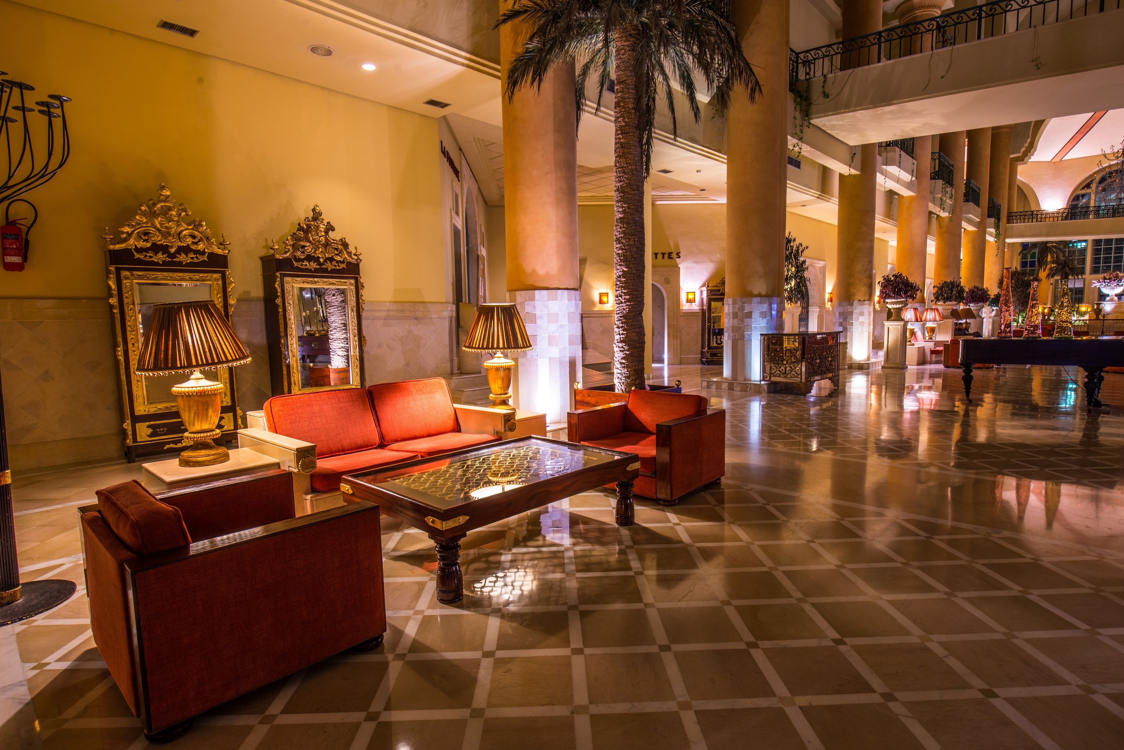 A spacious lobby with a checkered floor, red sofas, ornate lamps, and a large palm tree.