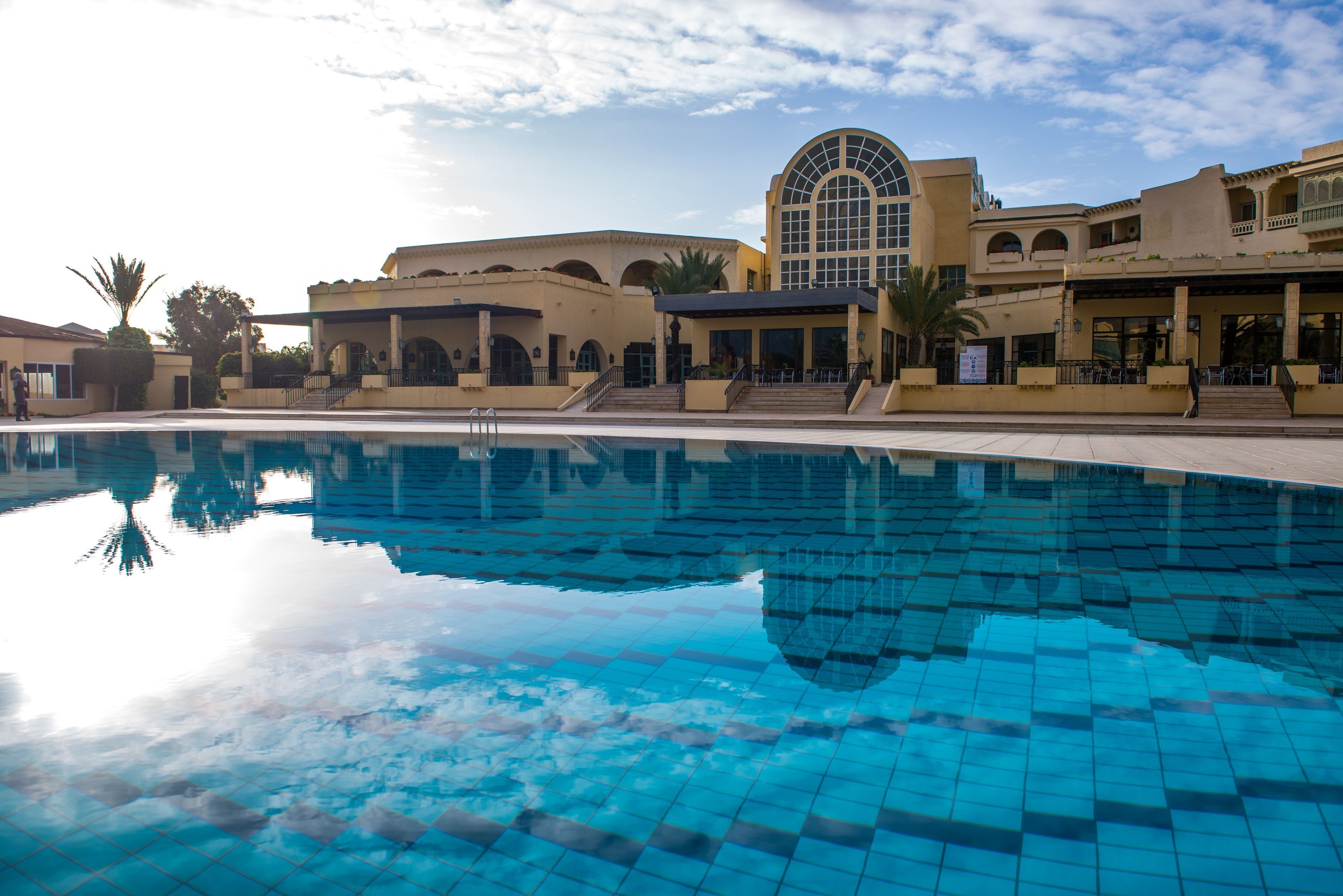 A pool area with a clear blue pool, a large arched window, and a building with multiple balconies and a covered patio.