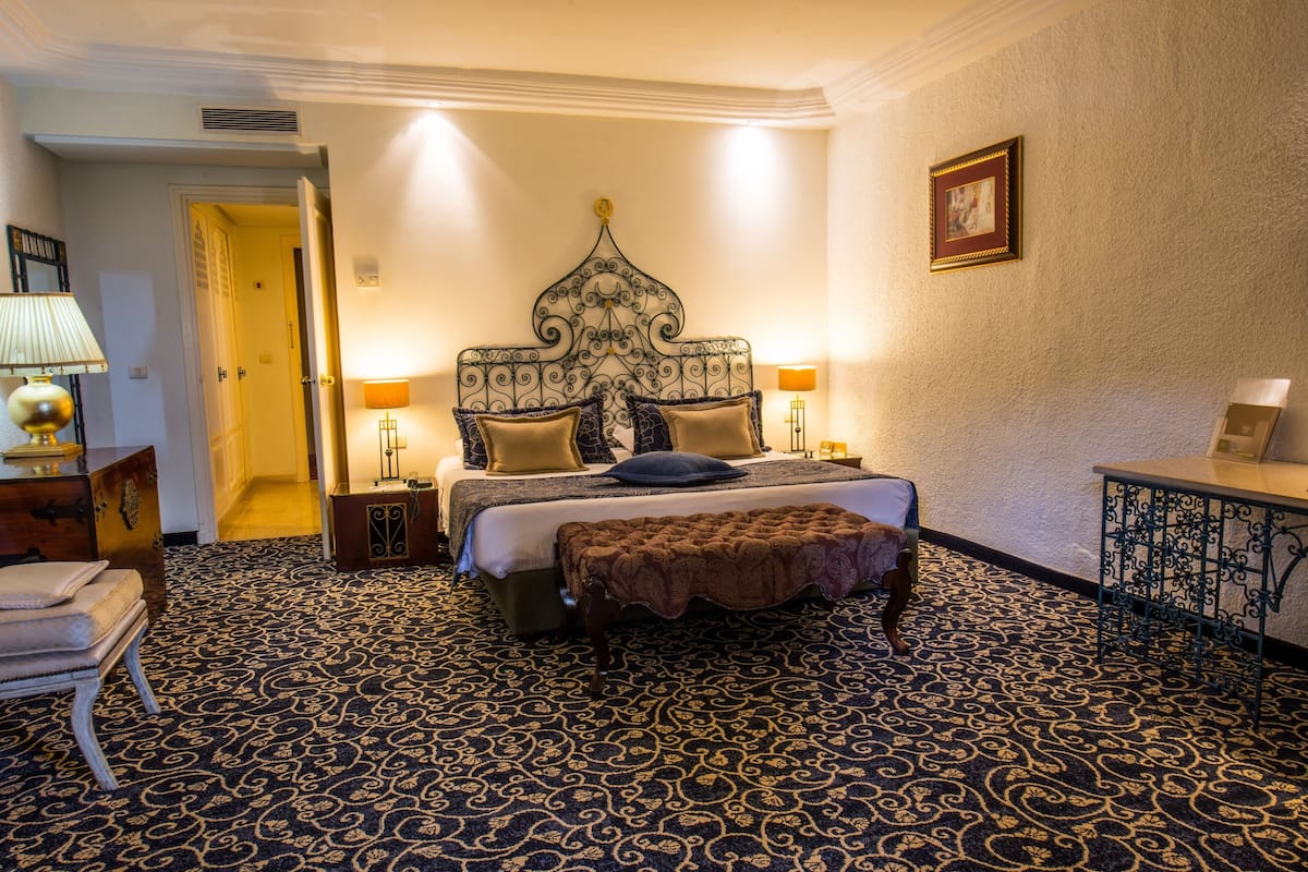 Habitación de hotel con una cama grande, una mesita de noche, una lámpara, un marco de foto y una alfombra estampada.