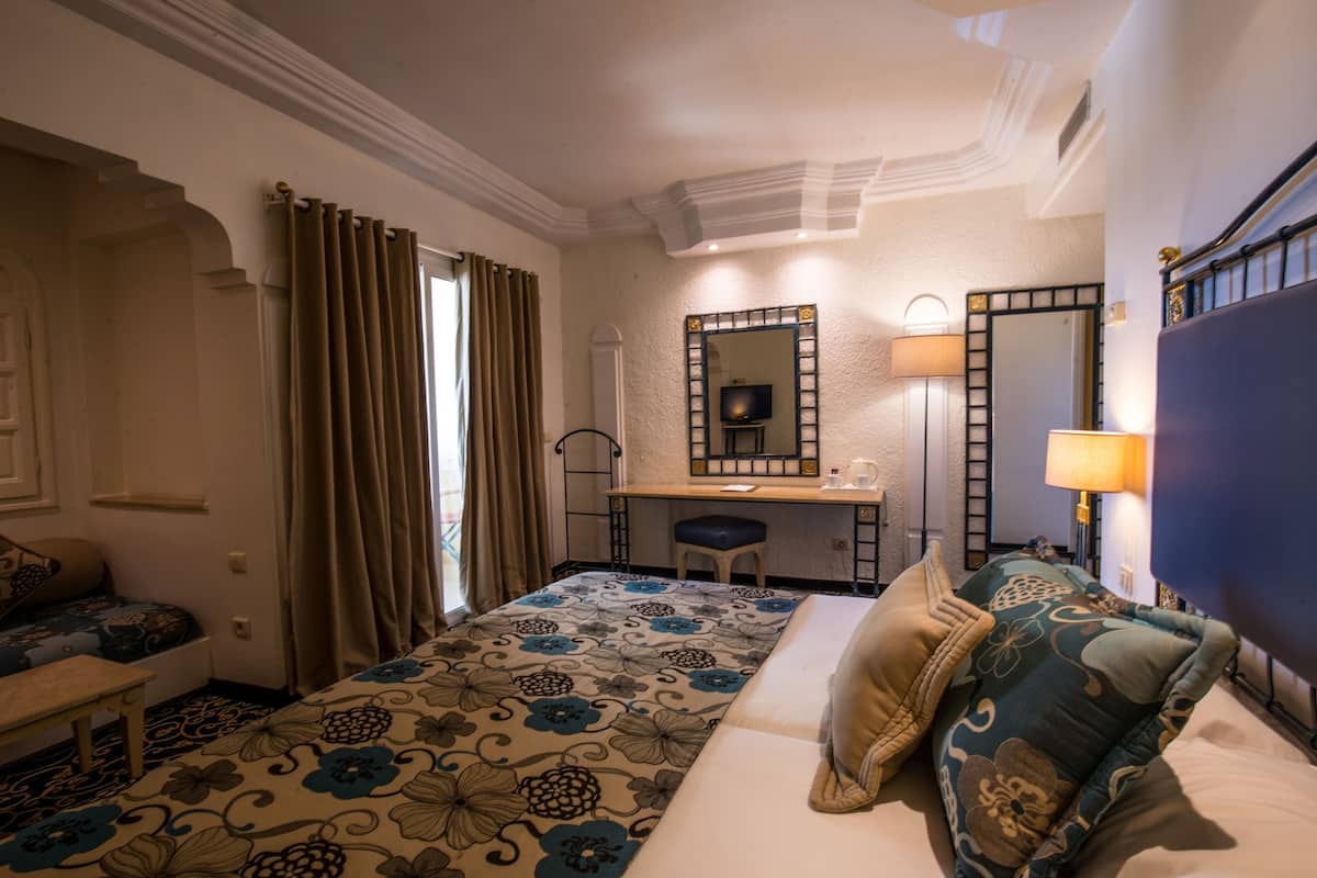 Habitación de hotel con cama, escritorio, silla, espejo y ventana con cortinas.