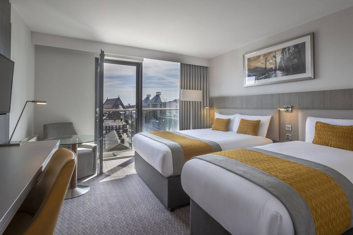 A hotel room with two beds, a desk, a chair, a lamp, and a view of the city through a large window.
