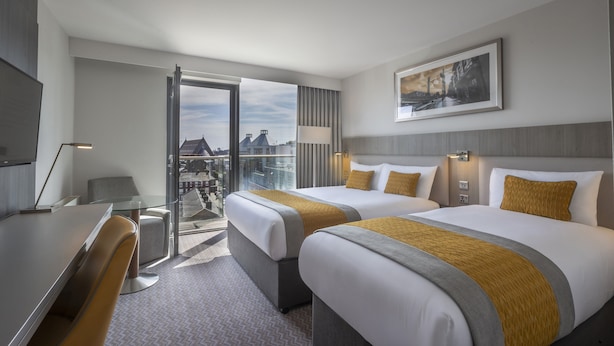 A hotel room with two beds, a desk, a chair, a lamp, and a view of the city through a large window.