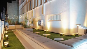 Un espacio exterior moderno con una larga banca, pequeñas mesas redondas y luces colgantes.