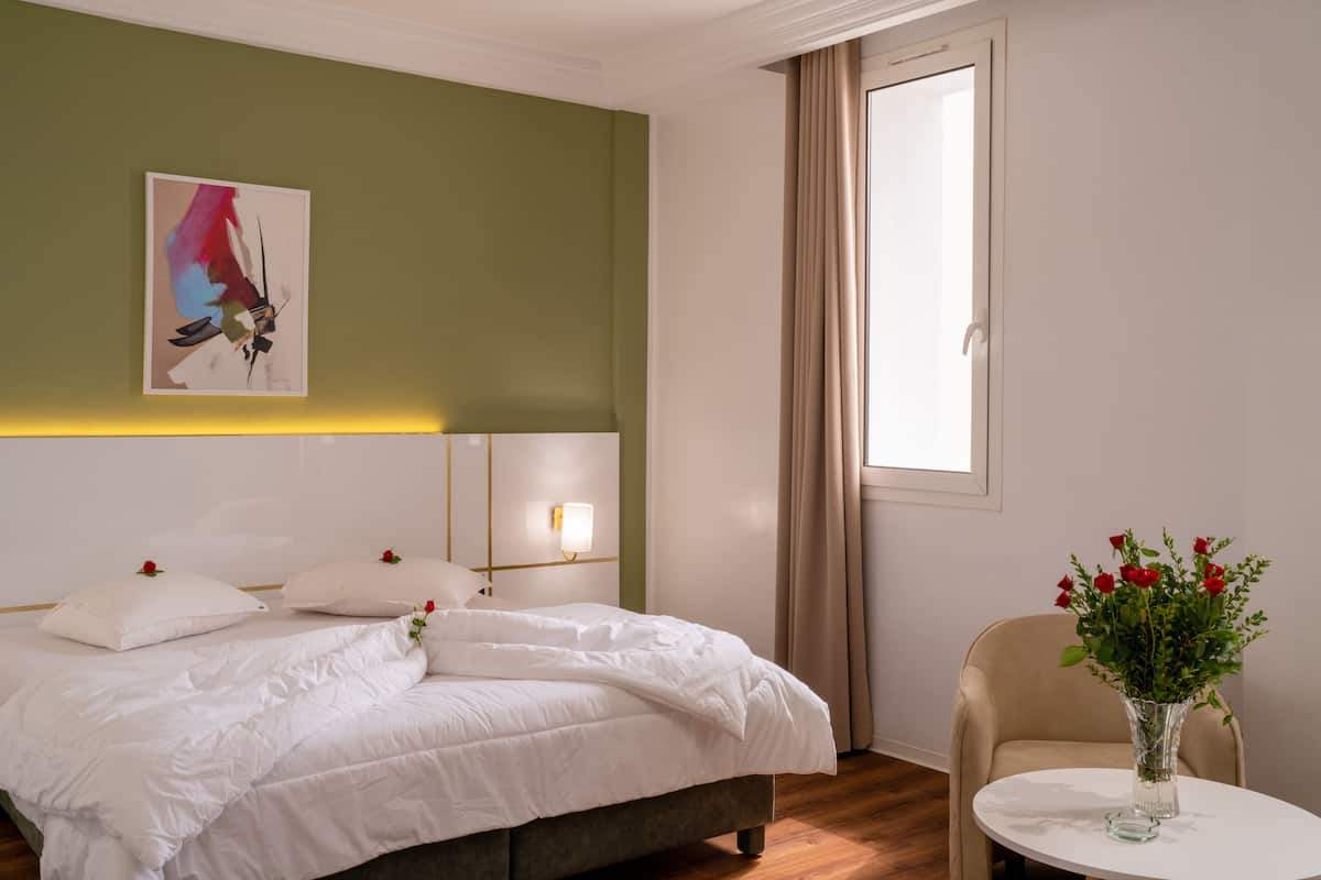 Habitación de hotel con cama, una silla, un jarrón con flores y un cuadro en la pared.