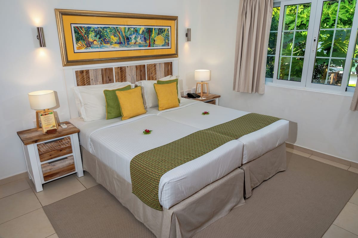 A hotel room with a large bed, wooden headboard, two bedside tables with lamps, a framed painting above the bed, and a window with curtains.
