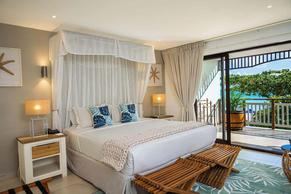 A bedroom with a large bed, a bedside table with a lamp, a wooden bench, and a view of the outdoors.