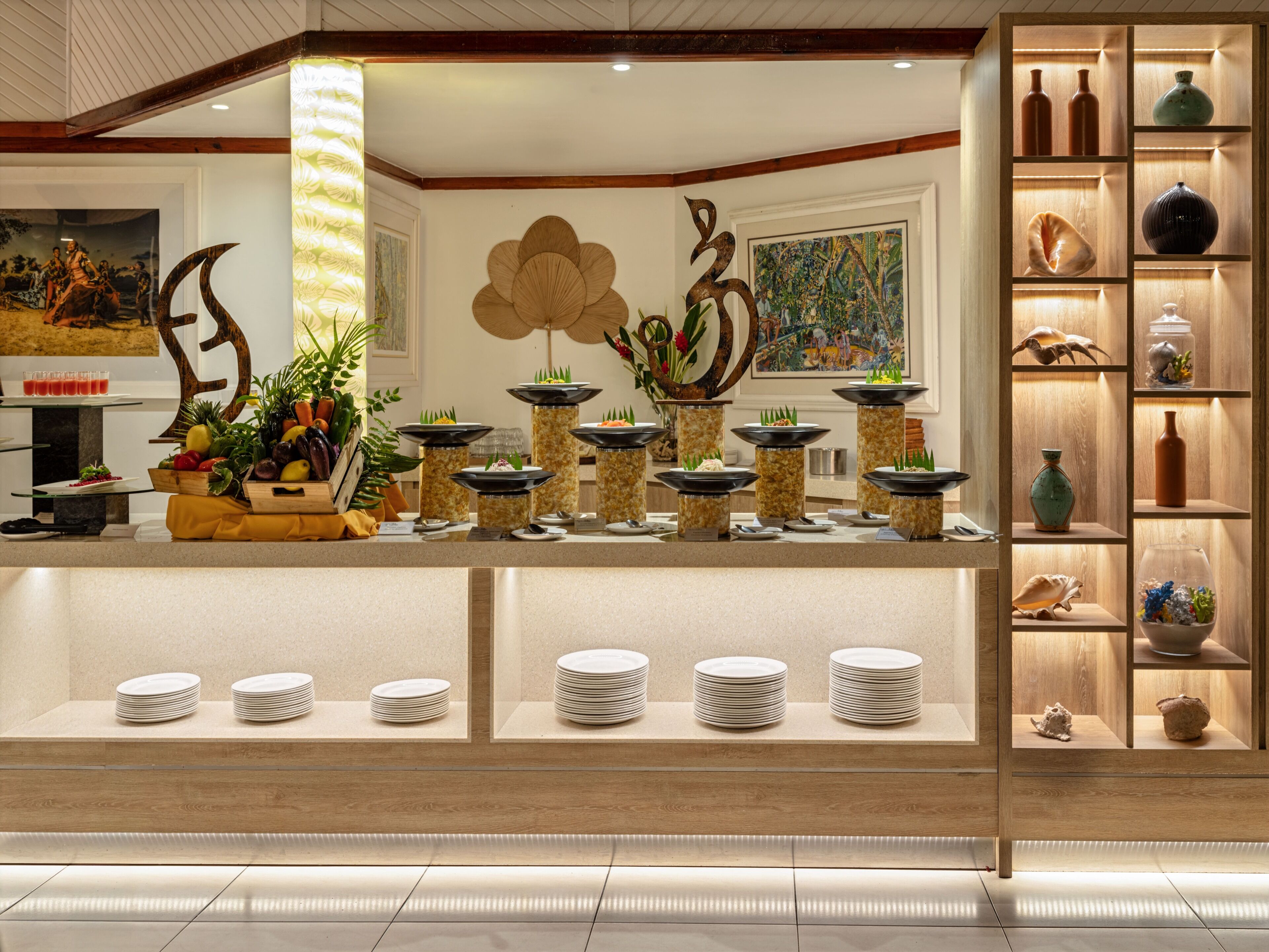 A hotel reception area with a central counter displaying a variety of fruits and vegetables, surrounded by decorative vases and a wall-mounted painting.