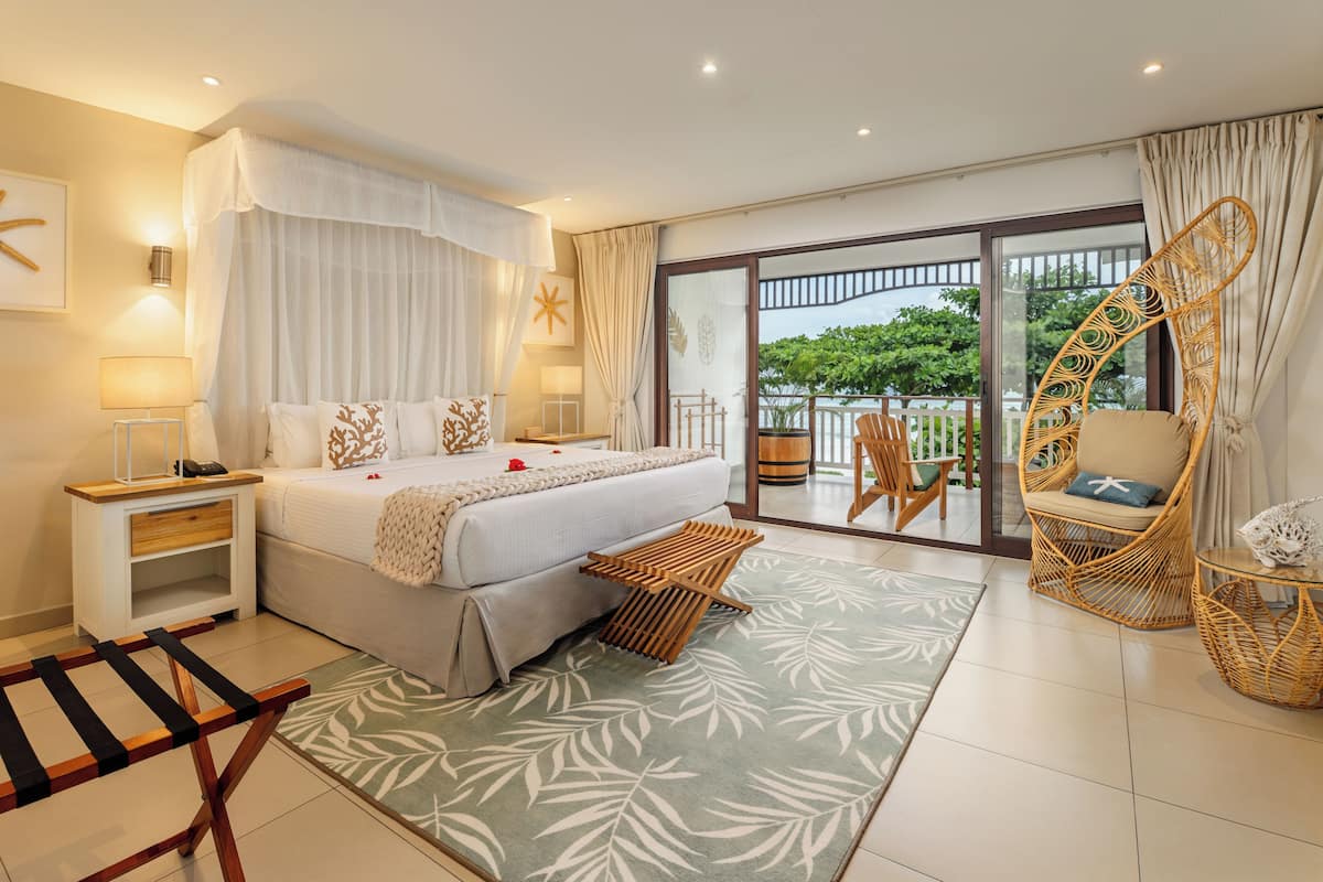 A bedroom with a bed, bedside tables, a nightstand, a chair, a small table, a balcony with a view, and a large window.