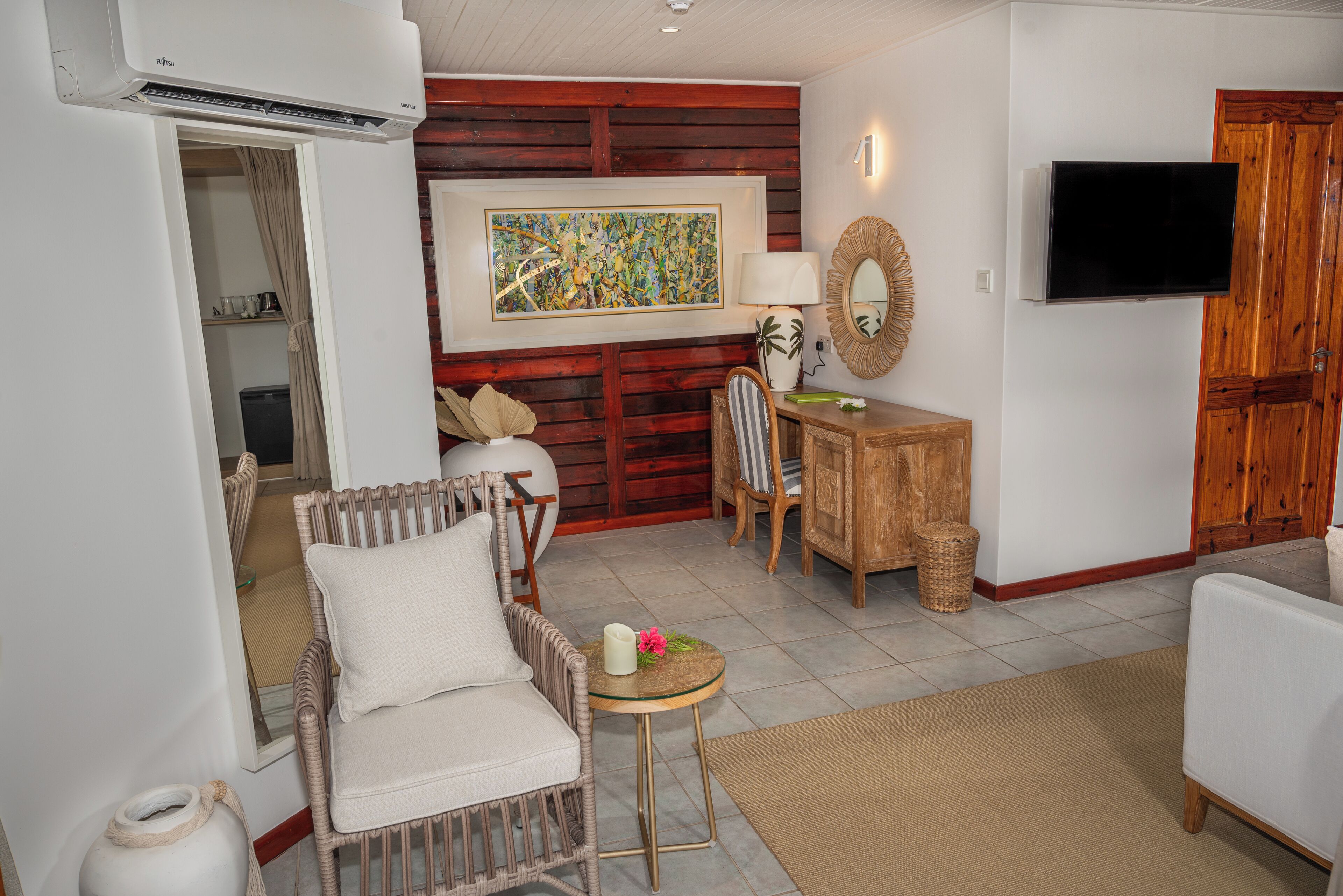 Una habitación con una silla de mimbre, una mesita redonda con una vela, un armario de madera y un televisor instalado en la pared.