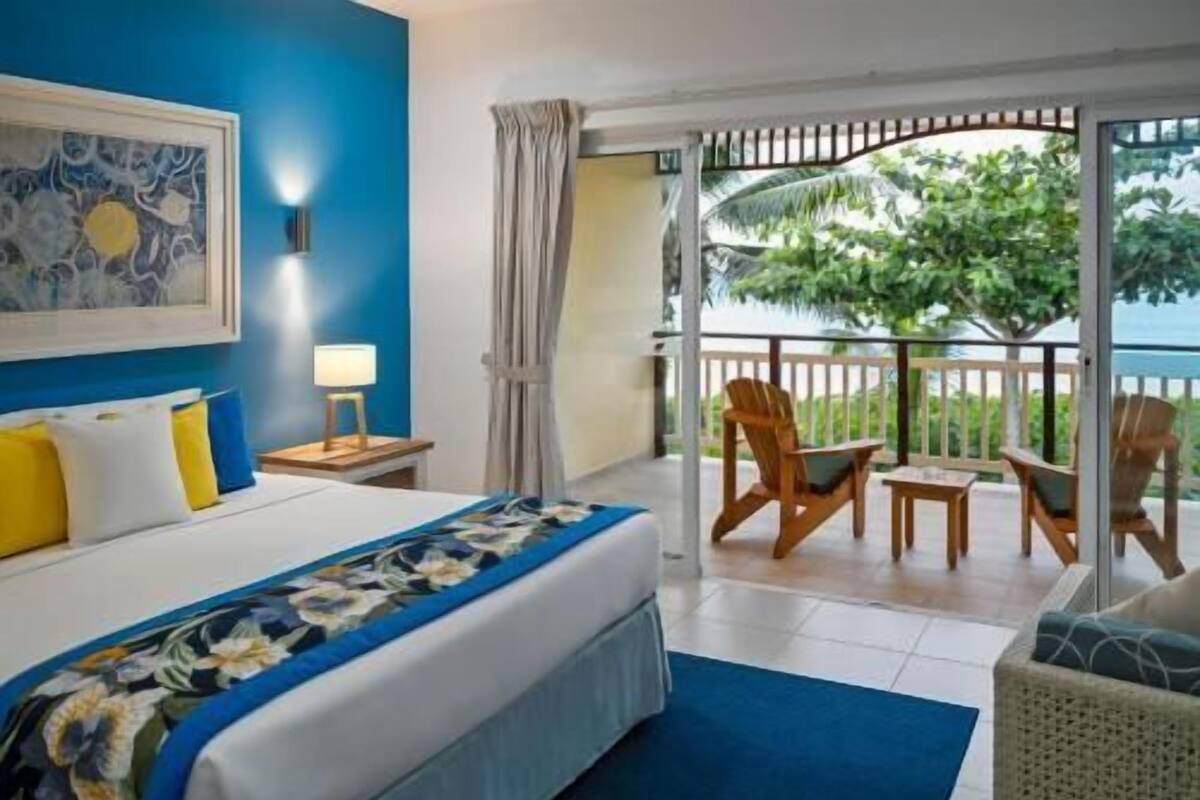 A hotel room with a bed, a nightstand, a lamp, a balcony with chairs, and a view of the outdoors.