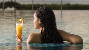 A person in a swimming pool holding a drink, with a bridge and greenery in the background. - Acajou Beach Resort (Praslin Island)