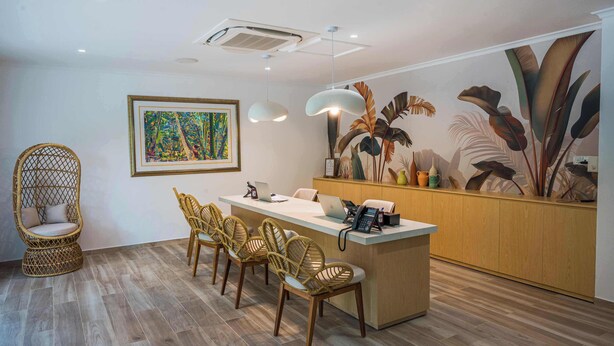 A modern reception area with a wooden reception desk, wicker chairs, a framed painting, and a tropical mural.