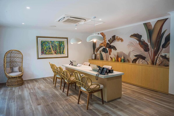A modern reception area with a wooden reception desk, wicker chairs, a framed painting, and a tropical mural. - Acajou Beach Resort (Praslin Island)