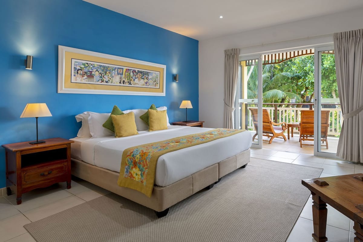 A bedroom with a large bed, two bedside tables, a nightstand, a window with curtains, and a view of greenery outside.