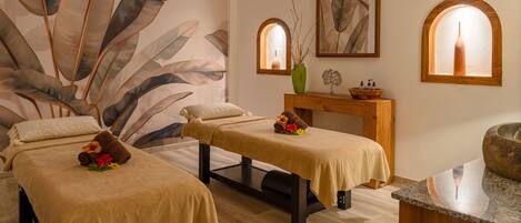 Un cuarto de spa con dos camillas de masaje, un banco de madera, un florero y una obra de arte enmarcada.