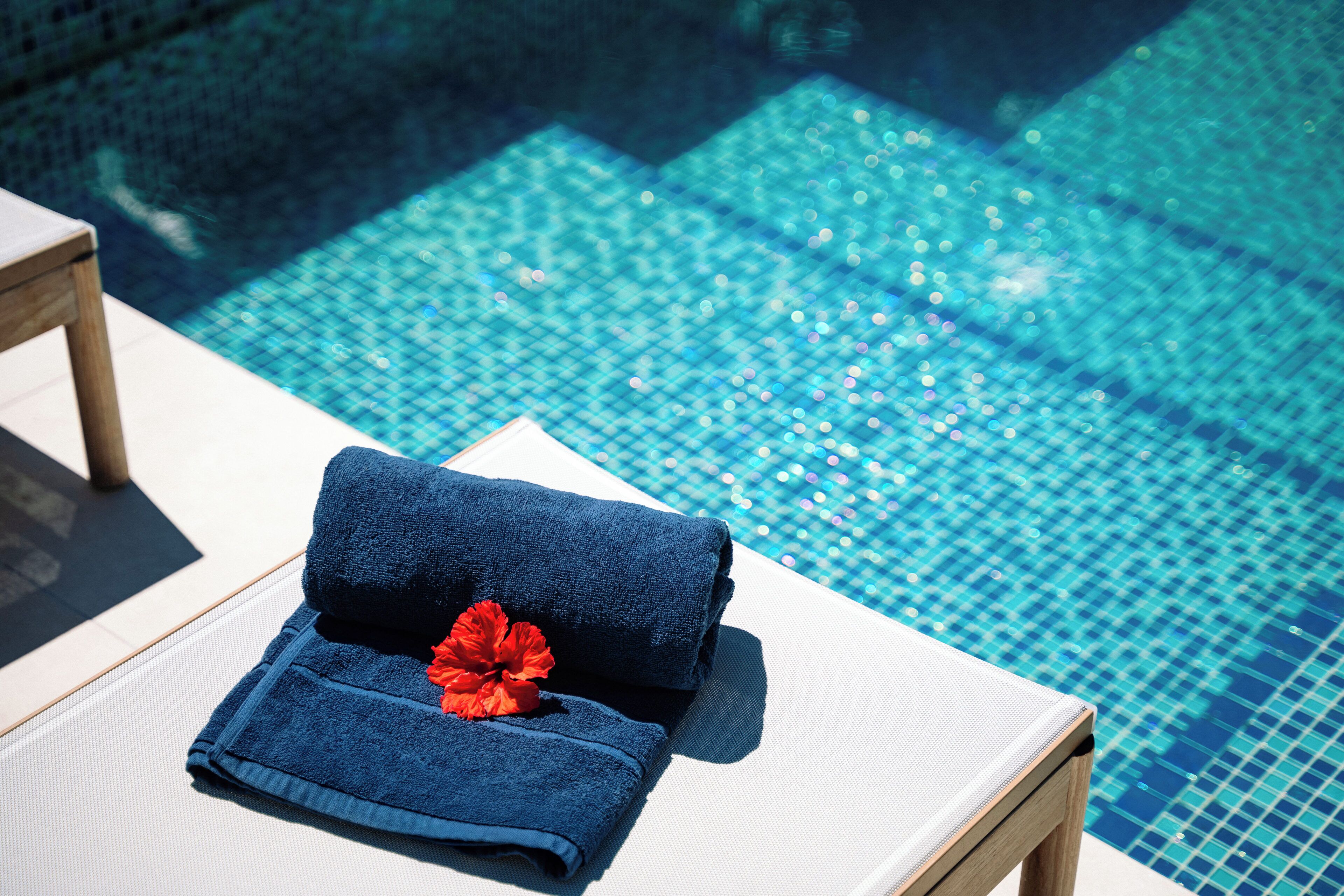 A poolside lounge area with a folded towel and a red flower on a white chair.