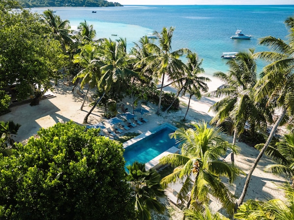 Un resort tropical con piscina, playa y embarcaciones al fondo.
