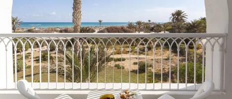A balcony with a white railing, white chairs, and a table with fruit and pastries. A beach with palm trees and a clear sky is visible in the background.