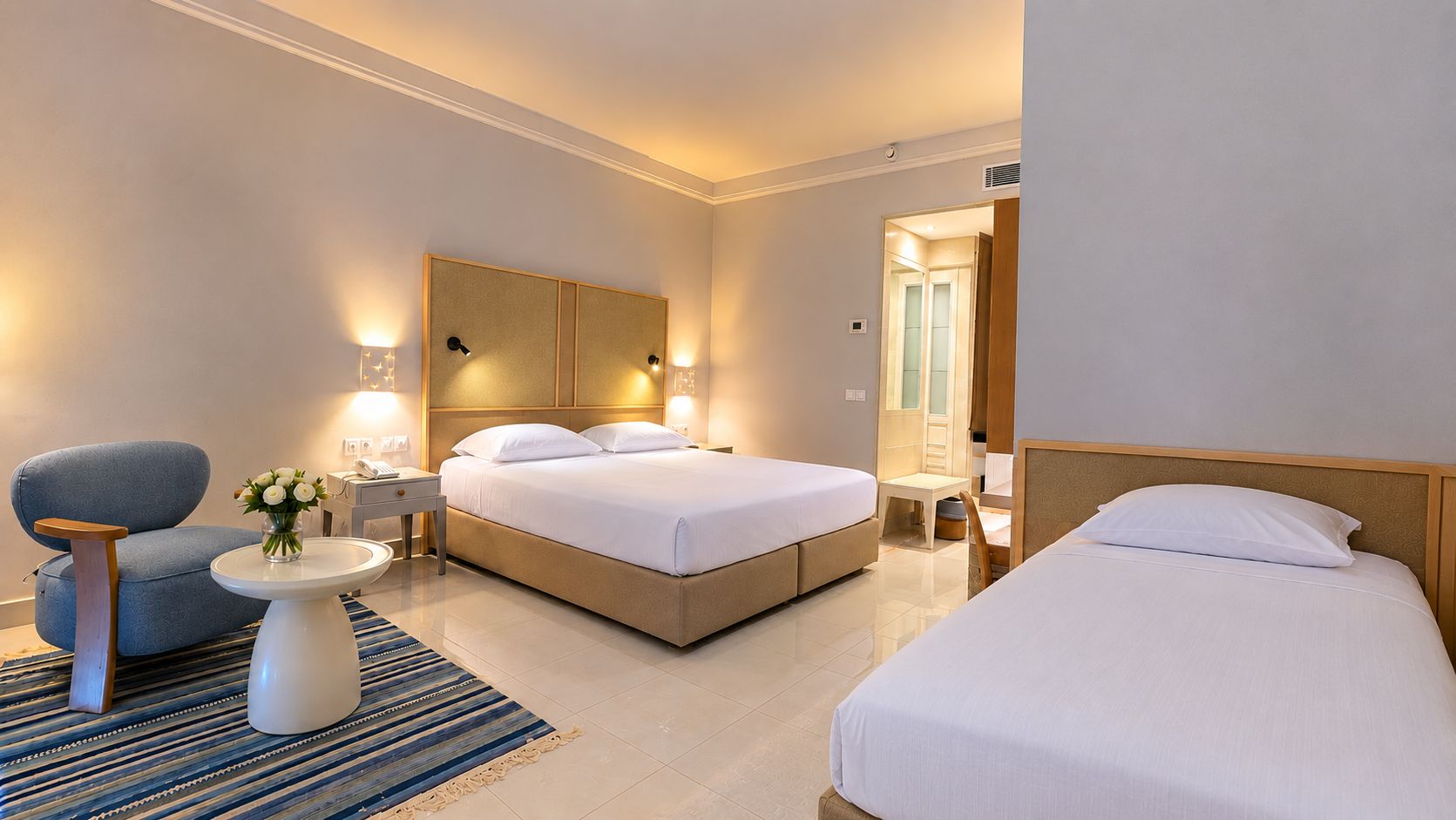 Ein Hotelzimmer mit Doppelbett, Einzelbett, einer Couch, einem kleinen Tisch mit Blumenvase und einem Nachttisch mit Lampe.