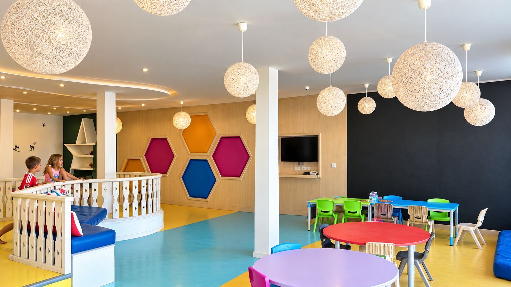 Zona de juegos infantiles con paneles hexagonales de colores, una estructura de juego blanca y un televisor de pantalla plana grande.