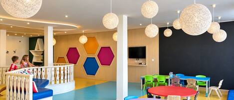 Zona de juegos infantiles con paneles hexagonales de colores, una estructura de juego blanca y un televisor de pantalla plana grande.