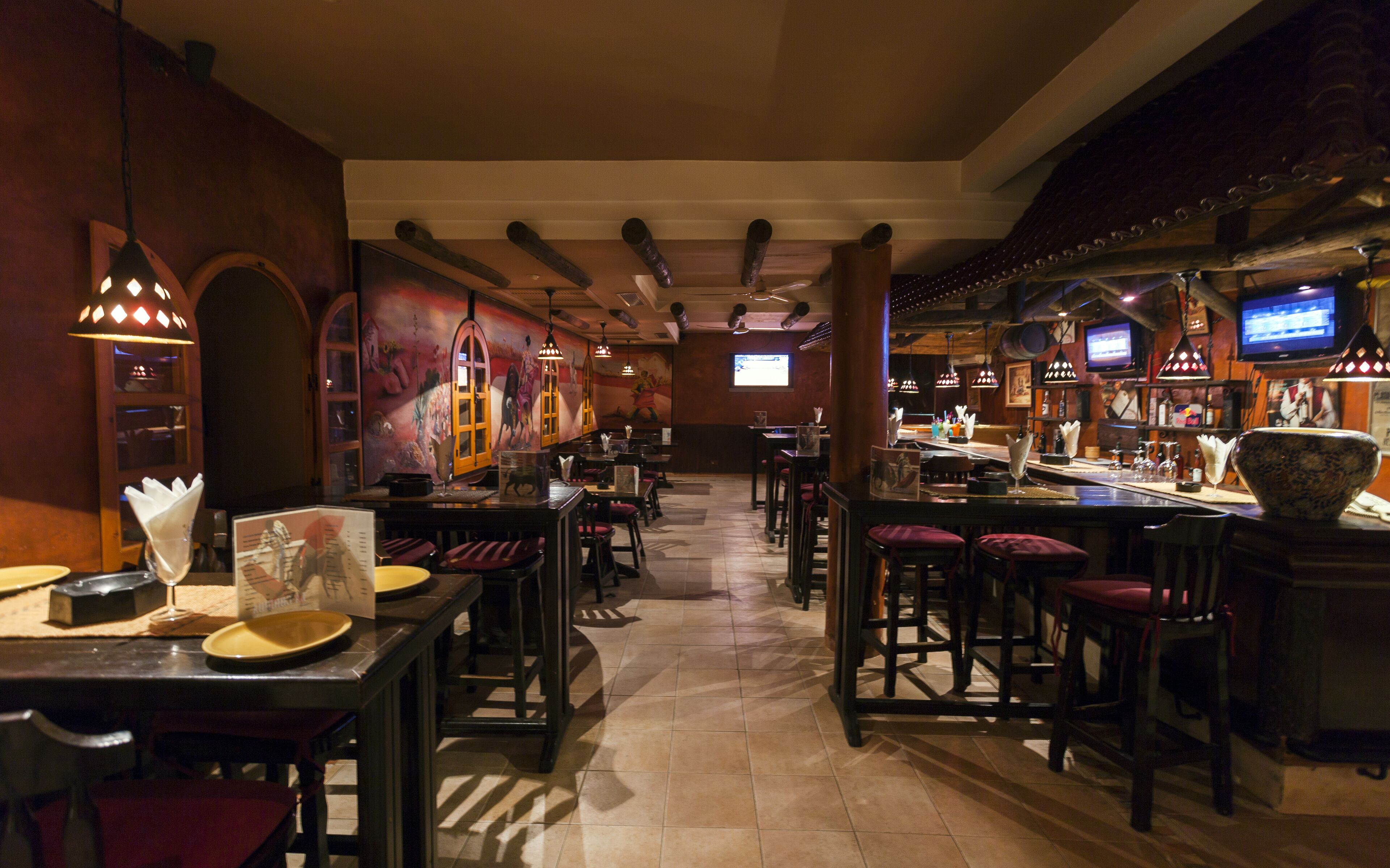 A restaurant interior with multiple tables and bar seating, a television screen, and a mural on the wall.