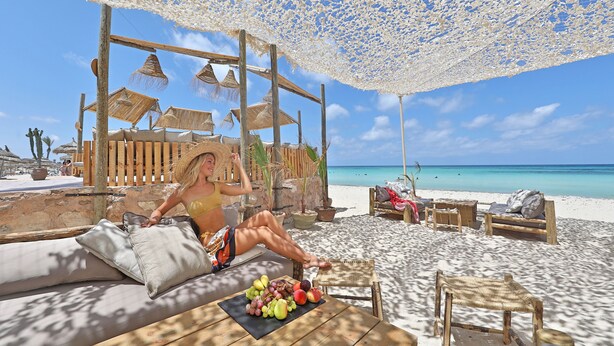 A beachside lounge with wooden furniture, a table with fruits, and a view of the ocean.