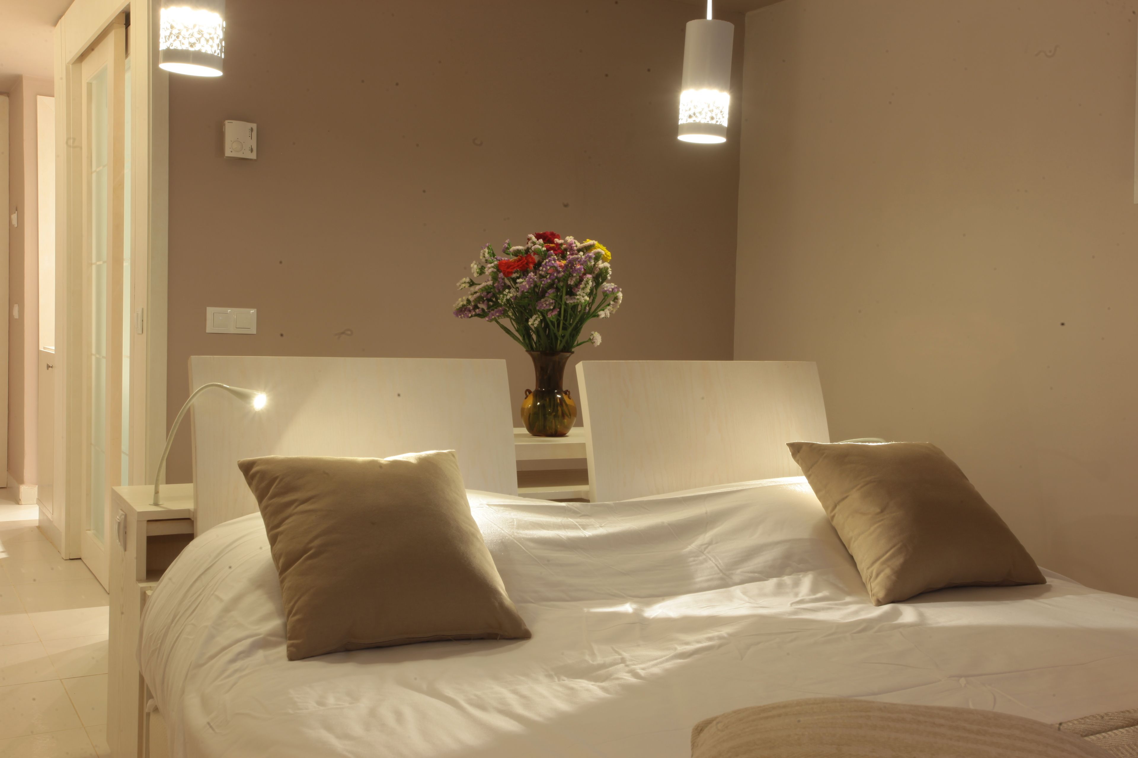 A neatly arranged bedroom with a bed, two bedside tables, a vase with flowers, and two pendant lights.