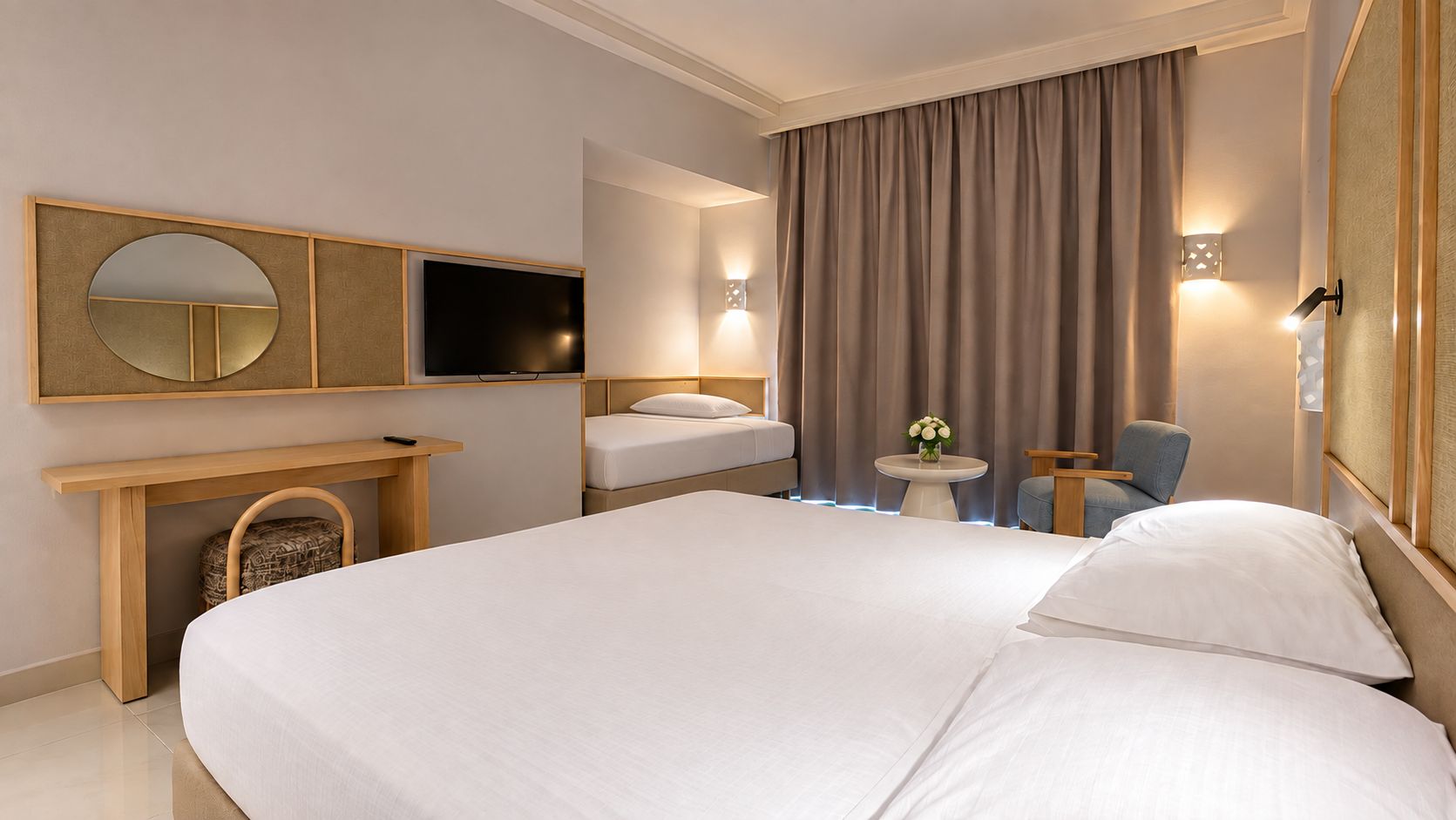 Habitación de hotel con una cama grande, televisor, escritorio con silla, una mesita con un jarrón de flores y ventana con cortinas.
