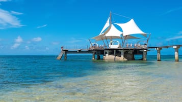 Un muelle que se adentra en el océano, con una zona de estar cubierta y una escalera que baja hacia el agua.