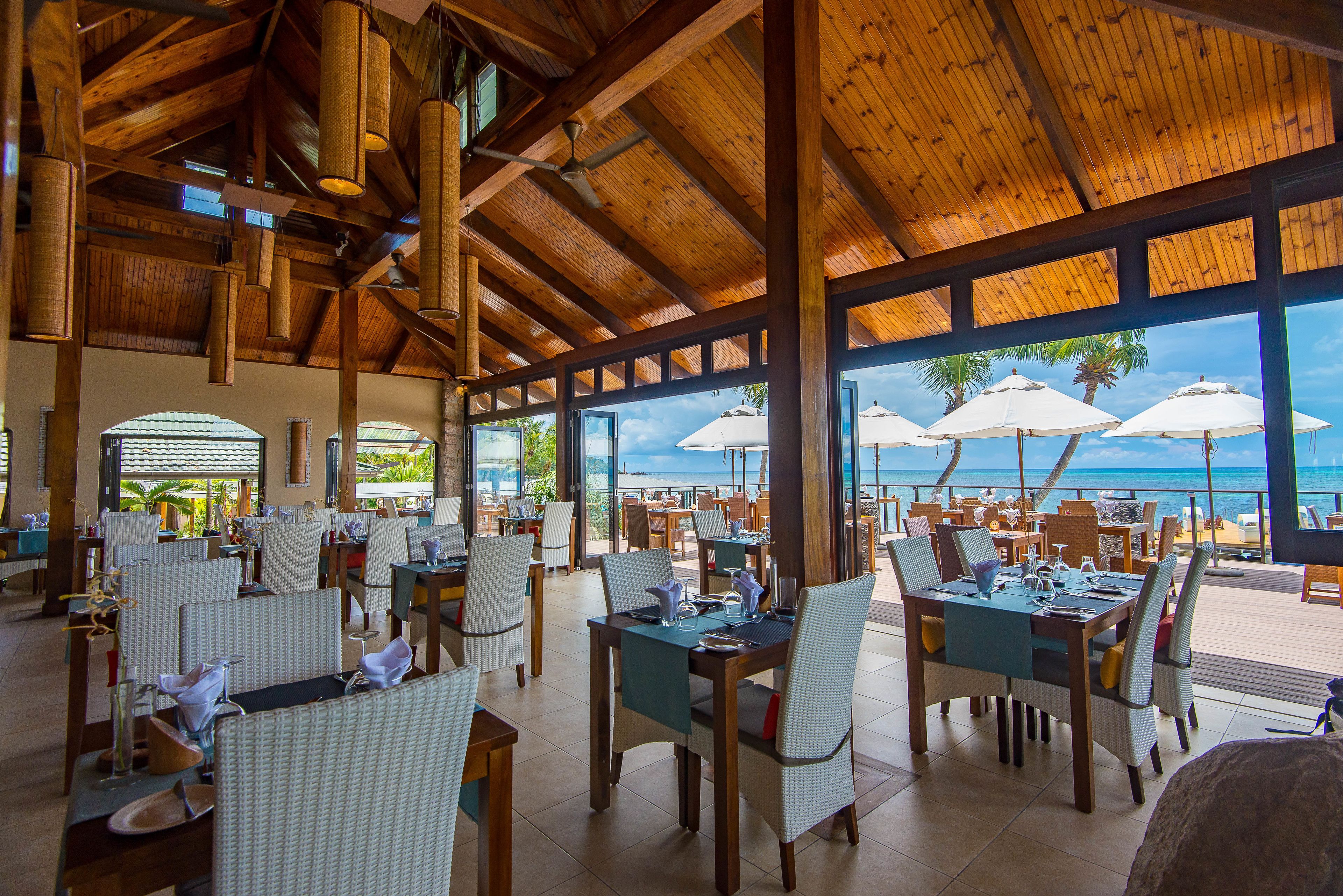 Um restaurante amplo com vigas de madeira e janelas grandes com vista para a praia. Mesas montadas com cadeiras e guarda-sóis.