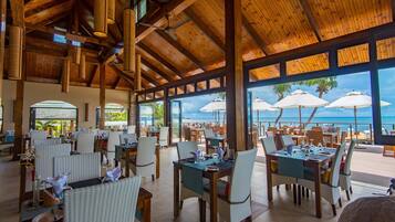 Um restaurante amplo com vigas de madeira e janelas grandes com vista para a praia. Mesas montadas com cadeiras e guarda-sóis.