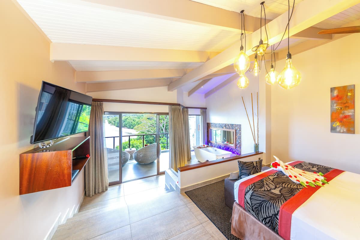 Een moderne hotelkamer met een groot bed, een flatscreen televisie bevestigd aan een houten plank, een balkon met rieten stoelen en een groot raam met gordijnen.