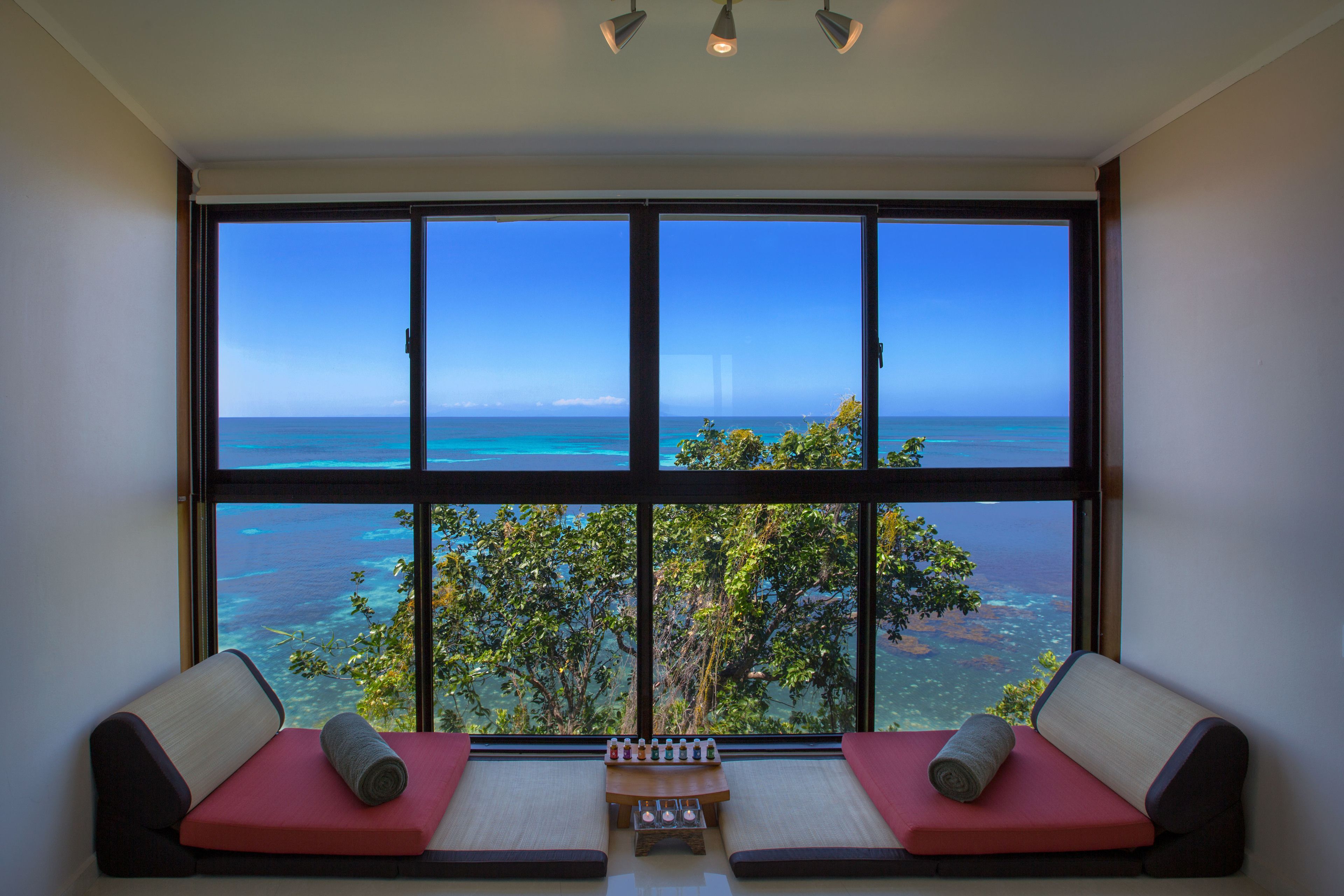 A room with a large window offering a view of the ocean and a tree. There are two red cushioned seats with pillows.