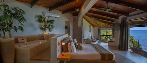 A spacious bedroom with a large bed, a sitting area with a bathtub, and a view of the ocean.