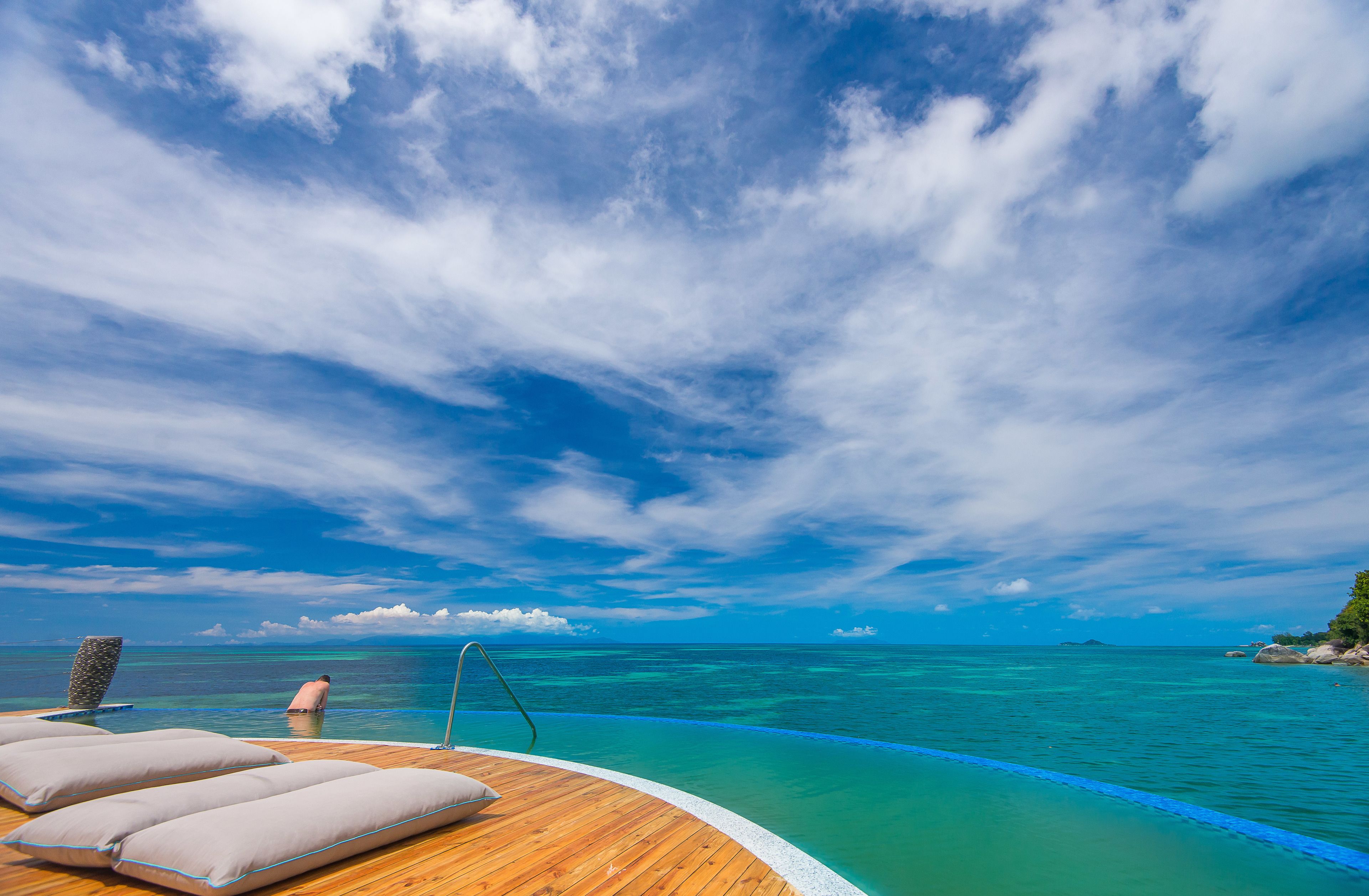 A wooden deck with lounge chairs and a pool overlooking turquoise waters and a clear sky.