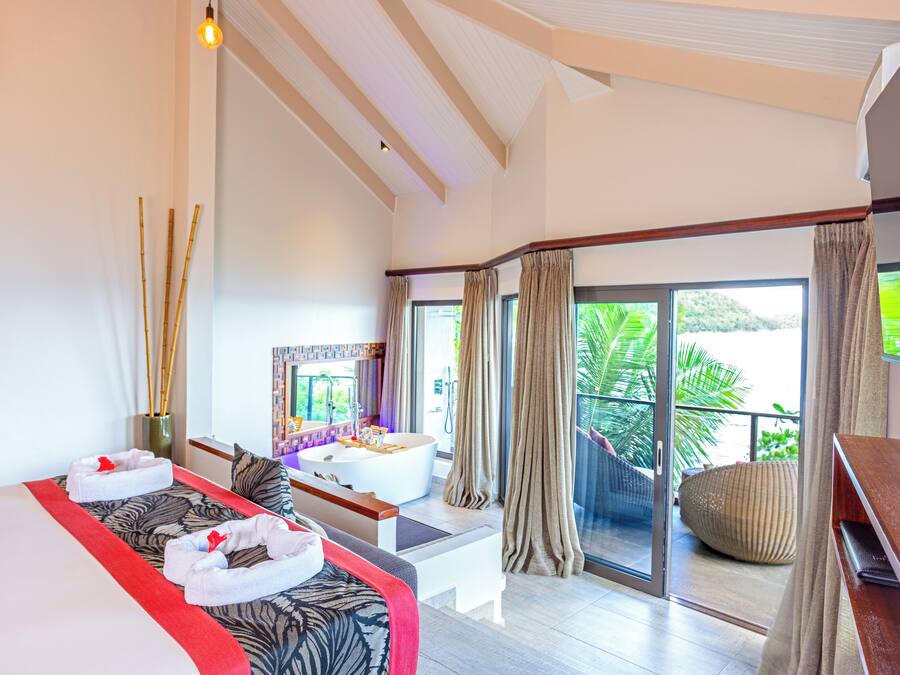 A modern hotel room with a large bed, a sitting area, a bathtub, and a balcony with a view of greenery.