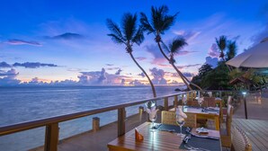 Een terras van een restaurant met tafels en stoelen, uitkijkend op de oceaan bij zonsondergang.