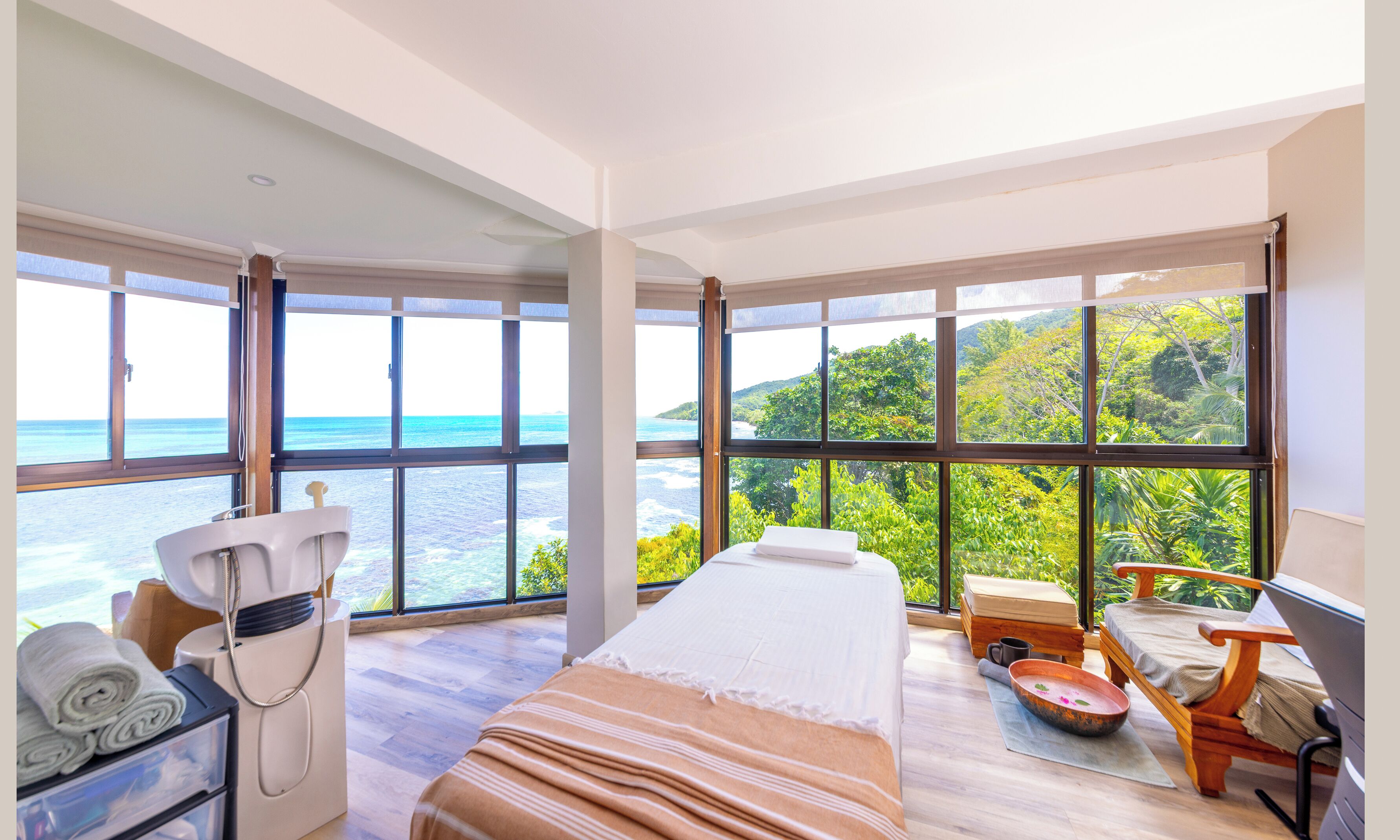Una habitación con cama, un sillón de masaje, una mesita con un cuenco y ventanas amplias con vistas al mar y al follaje.