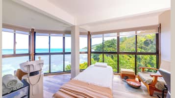 Una habitación con cama, un sillón de masaje, una mesita con un cuenco y ventanas amplias con vistas al mar y al follaje.