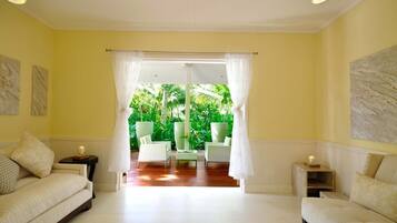 A room with a wooden floor, two white sofas, a small table, and chairs. There is a view of greenery outside through a window with sheer curtains.