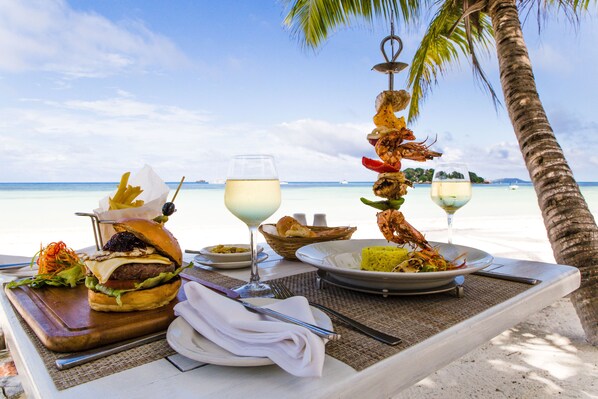 A dining table with a meal, including a burger, shrimp skewer, and rice, set on a beach with a palm tree and ocean view. - Paradise Sun (Praslin Island)