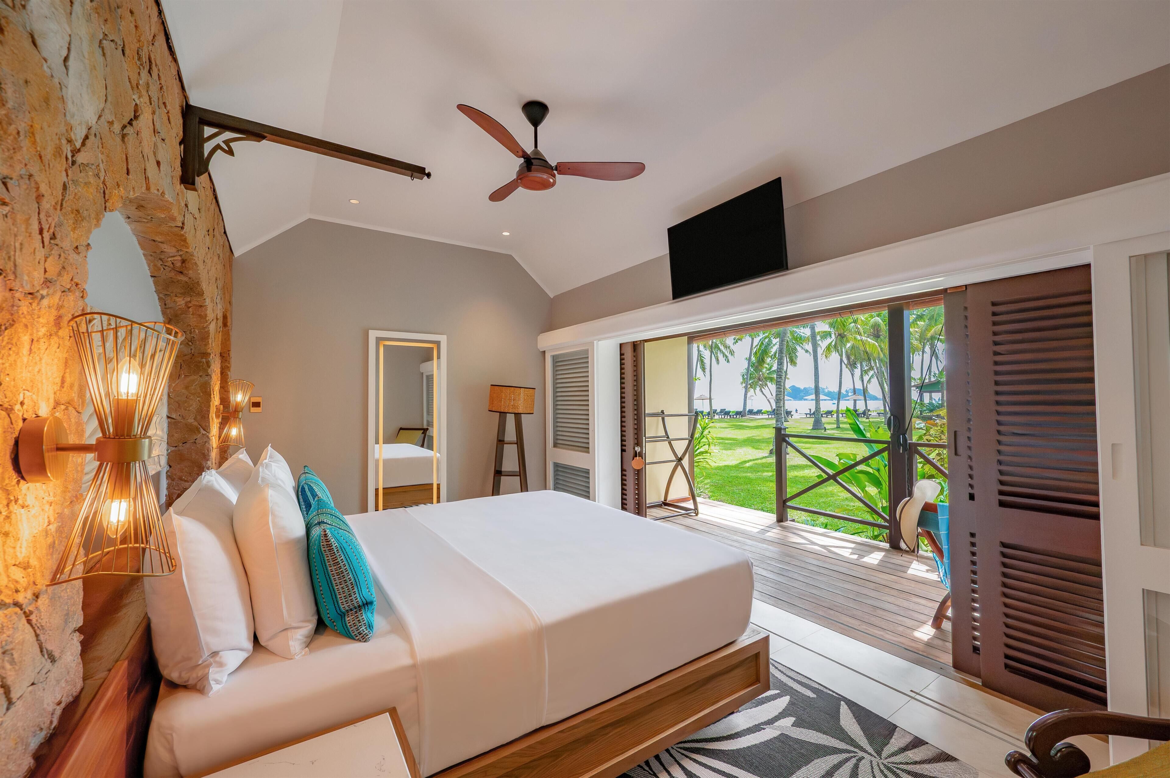 A bedroom with a bed, a ceiling fan, a TV, and a view of a green landscape through a sliding door.