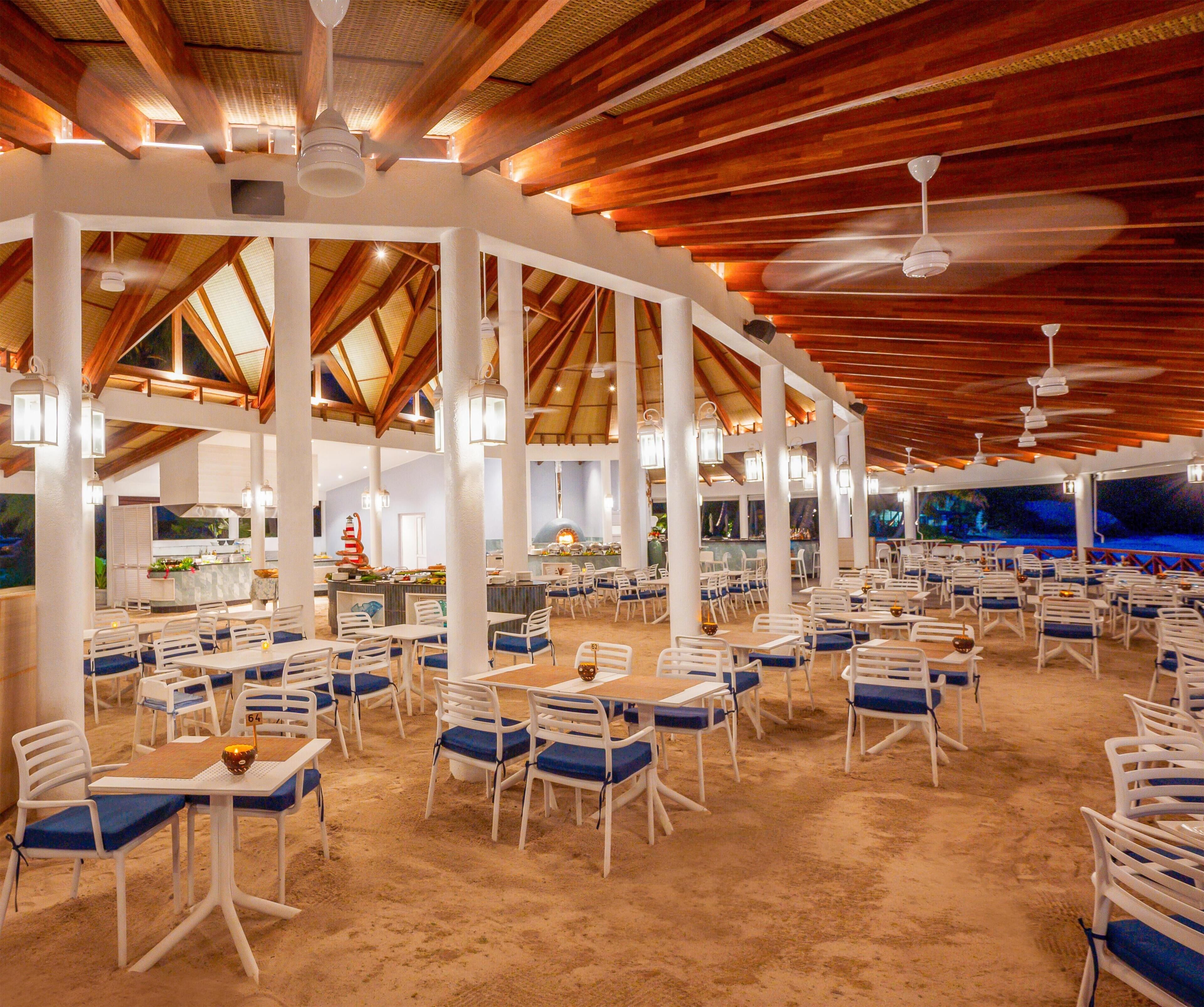 Un vaste espace de restauration intérieur avec des tables blanches et des chaises rembourrées bleues, des poutres en bois au plafond et des ventilateurs de plafond.