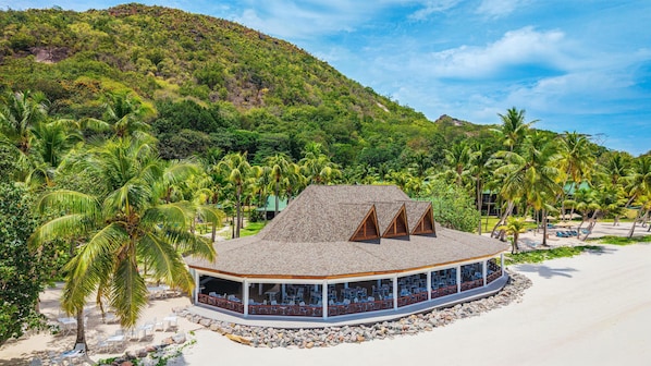Un resort frente a la playa con zona de comedor cubierta, asientos al aire libre y vistas a la playa y las montañas.