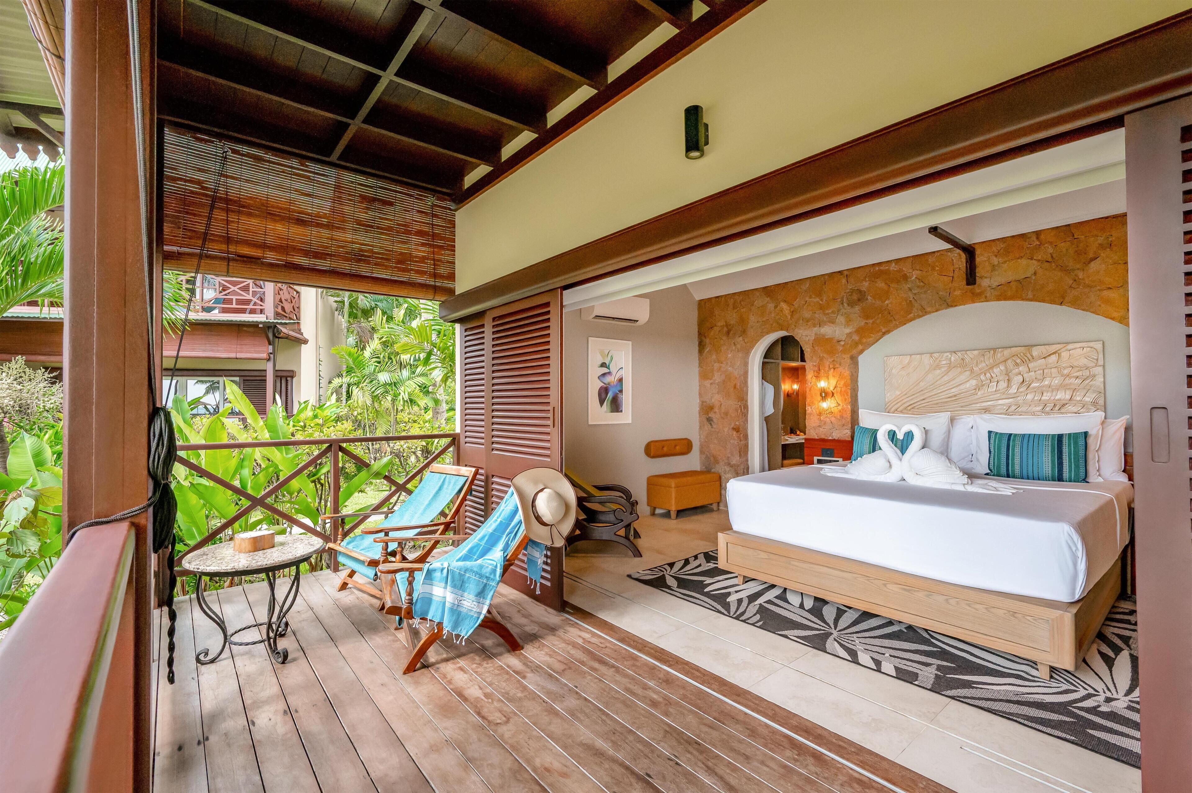 Un dormitorio con una cama grande, cabecera de madera, balcón con sillas y vista a zonas verdes.