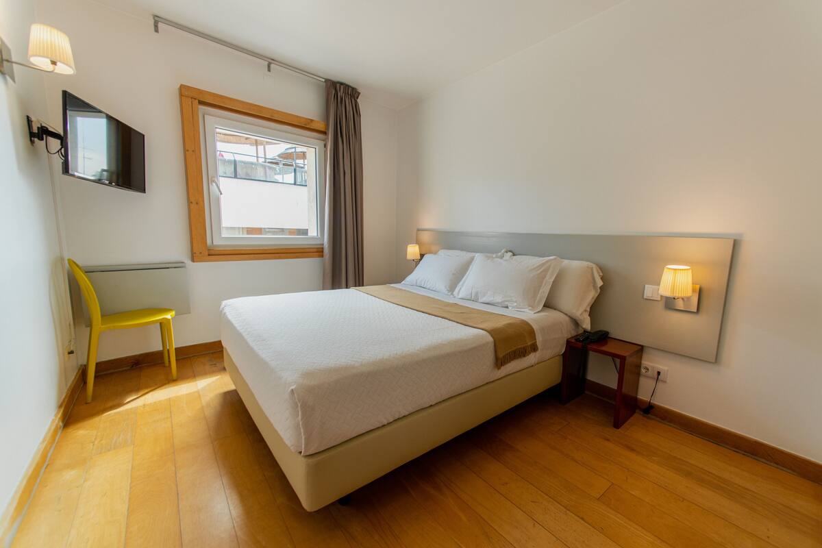 Una habitación de hotel con una cama, una silla, un televisor, una ventana y una mesita de noche.
