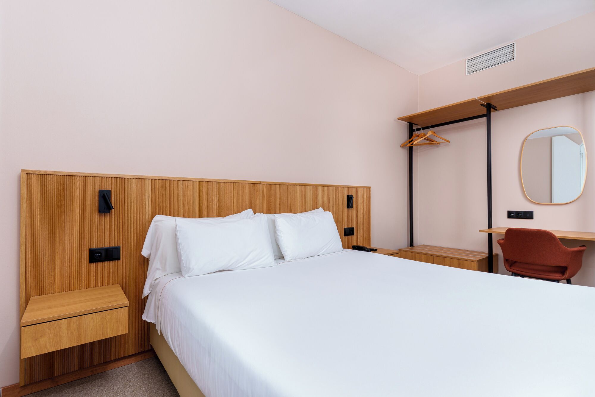 Een hotelkamer met een houten hoofdbord, een bed met witte lakens, een bureau met stoel, en een spiegel aan de muur.