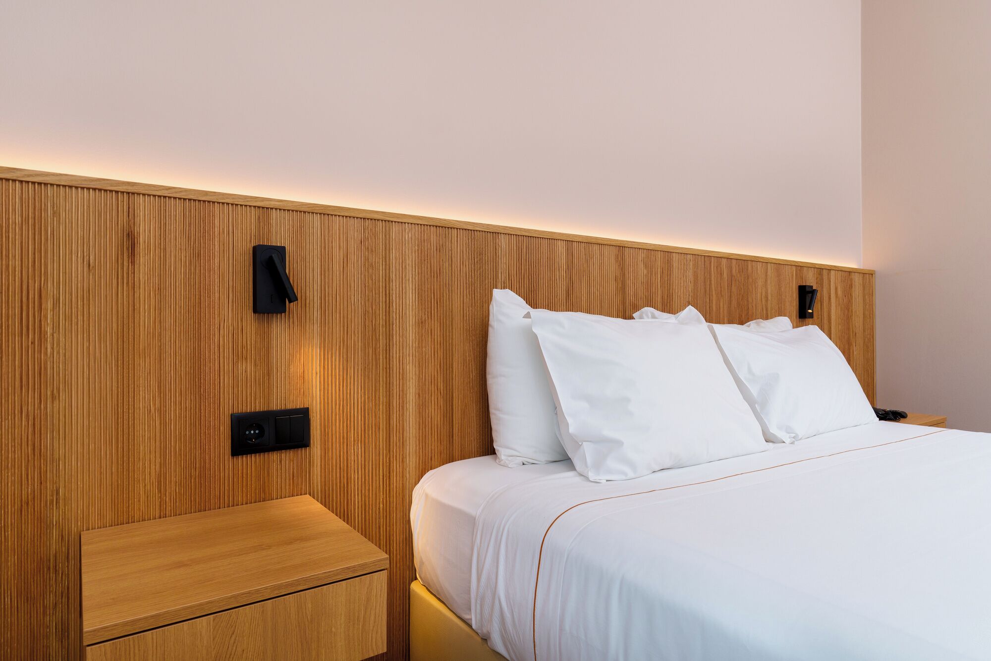 A hotel room with a wooden headboard, a bedside table, and a bed with white bedding.