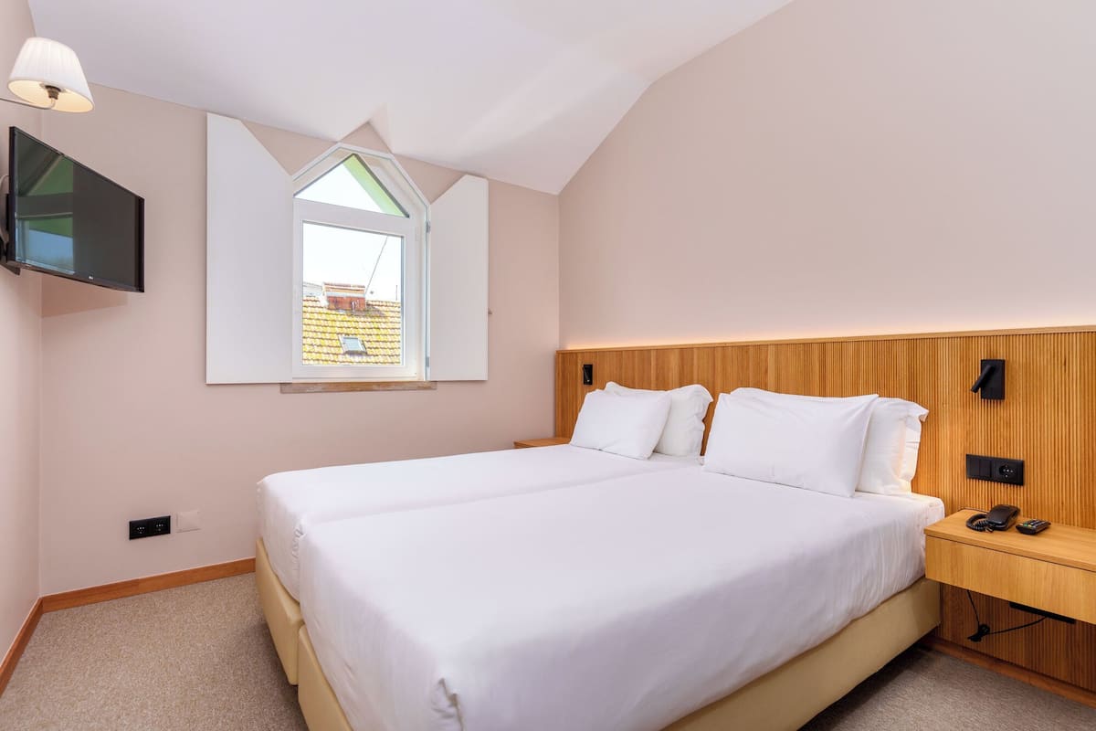 Un dormitorio con una cama, televisor montado en la pared, mesita de noche con un teléfono y una ventana con vistas al exterior.